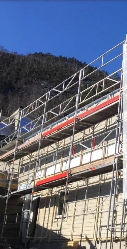 Fasadestillas montert rundt et trehus, med fjell og blå himmel bak